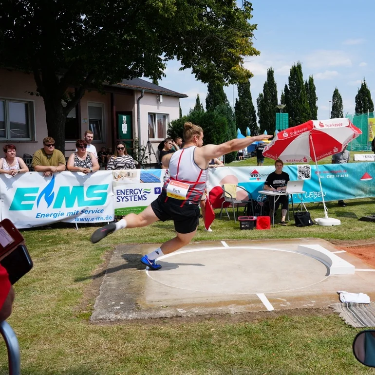 Ein Kugelstoßer in der Bewegung, die Kugel stoßend, aufgenommen. Aufnahme auf einem Sportplatz. An den Abgrenzungen stehen Menschen und beobachten die Szenerie.