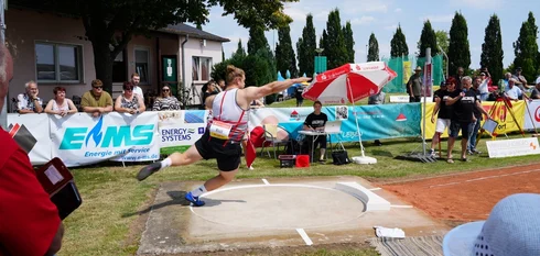 Ein Kugelstoßer in der Bewegung, die Kugel stoßend, aufgenommen. Aufnahme auf einem Sportplatz. An den Abgrenzungen stehen Menschen und beobachten die Szenerie.