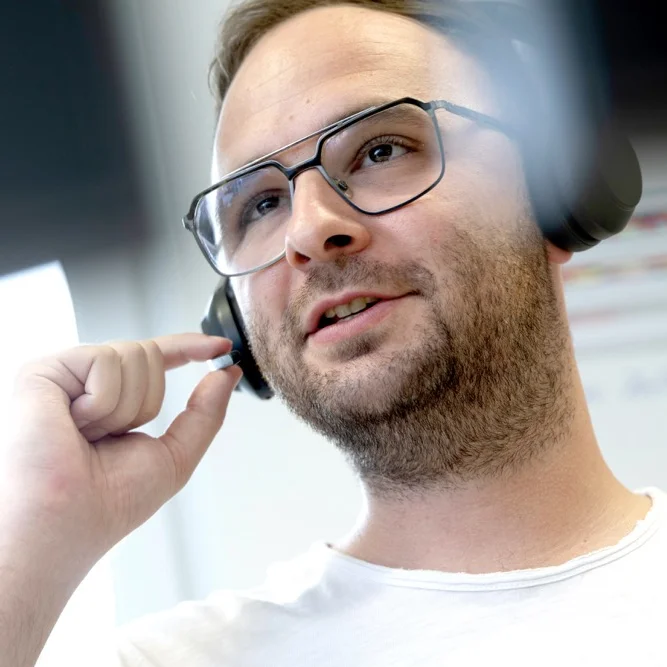 Ein bärtiger, junger Mann mit Brille wurde aus einer Perspektive von unten während eines Gesprächs über ein Headset fotografiert.