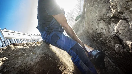 Ein Mann in einem blauen Arbeitsanzug platziert bei strahlendem Sonnenschein und blauem Himmel sorgfältig ein Rohr in einem vorbereiteten Baugraben.