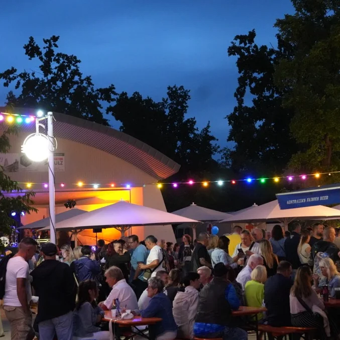 Viele Menschen sitzen am Abend auf Bierbänken an einem Getränkestand. Im Hintergrund eine beleuchtete Bühne