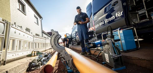 Zwei Männer in blauen EMS-Arbeitssachen stehen auf einer Baustelle, während rechts der offene EMS-Transporter zu sehen ist; der eine Mann steht davor und schaut auf ein Tablet, während der andere ein Rohr in ein unterstützendes Arbeitsgerät platziert.