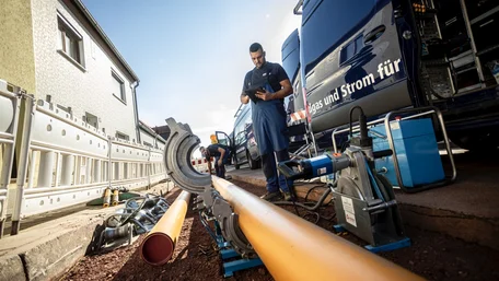 Zwei Männer in blauen EMS-Arbeitssachen stehen auf einer Baustelle, während rechts der offene EMS-Transporter zu sehen ist; der eine Mann steht davor und schaut auf ein Tablet, während der andere ein Rohr in ein unterstützendes Arbeitsgerät platziert.