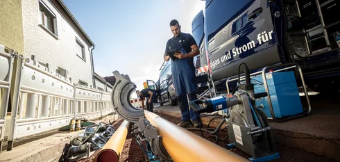 Zwei Männer in blauen EMS-Arbeitssachen stehen auf einer Baustelle, während rechts der offene EMS-Transporter zu sehen ist; der eine Mann steht davor und schaut auf ein Tablet, während der andere ein Rohr in ein unterstützendes Arbeitsgerät platziert.