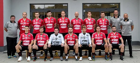 Mannschaftsfoto einer Handballmannschaft mit 14 Spielern in roten Trikots und 2 Trainern in grauen Shirts vor einem Gebäude