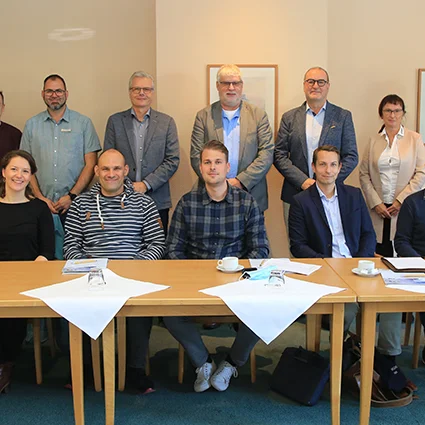 Gruppenfoto von 15 Personen, stehend und sitzend, in einem Konferenzraum hinter einem gedeckten Tisch.