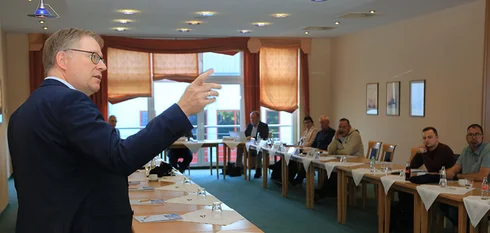 Ein Mann hält einen Vortrag vor einer Gruppe sitzender Menschen in einem Konferenzraum mit großen Fenstern.