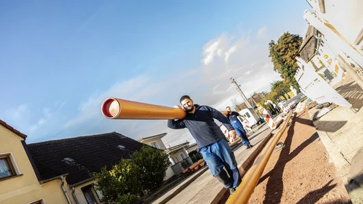 Zwei Männer in blauer EMS-Arbeitskleidung tragen bei gutem Wetter gemeinsam ein oranges Rohr über eine Baustelle.