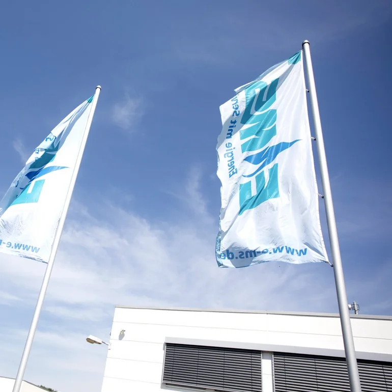 Drei weiße, von unten betrachtete, EMS-gebrandete Flaggen wehen im Wind bei blauem Himmel.