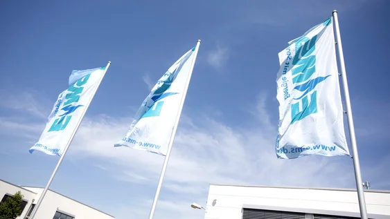 Drei weiße, von unten betrachtete, EMS-gebrandete Flaggen wehen im Wind bei blauem Himmel.