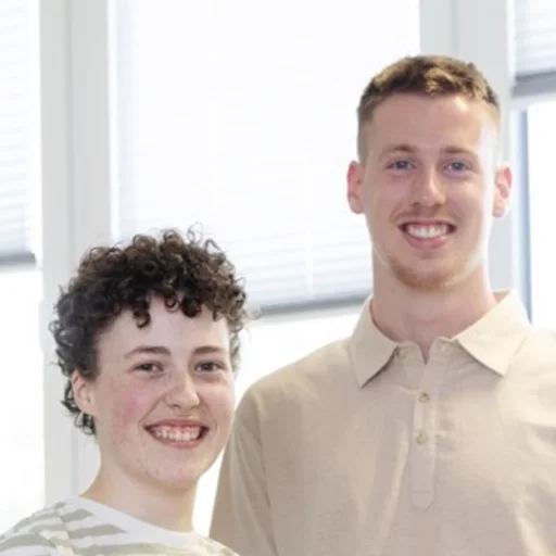 Ein Mann und eine Frau stehen als Team frontal zur Kamera, selbstbewusst und lachend, an einem Schreibtisch vor einem großen Fenster, wobei sie die Arme vor der Brust verschränkt und er sich mit einem Arm auf dem Schreibtisch hinter ihr abstützt.