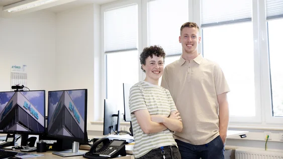 Ein Mann und eine Frau stehen als Team frontal zur Kamera, selbstbewusst und lachend, an einem Schreibtisch vor einem großen Fenster, wobei sie die Arme vor der Brust verschränkt und er sich mit einem Arm auf dem Schreibtisch hinter ihr abstützt.