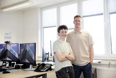 Ein Mann und eine Frau stehen als Team frontal zur Kamera, selbstbewusst und lachend, an einem Schreibtisch vor einem großen Fenster, wobei sie die Arme vor der Brust verschränkt und er sich mit einem Arm auf dem Schreibtisch hinter ihr abstützt.