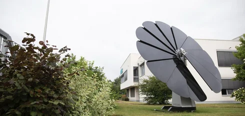 Die aufgefahrene große E-Solarblume auf der Grünfläche, des großen weißen EMS-Gebäudes, bei bedecktem Himmel.