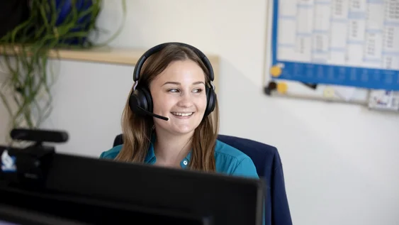 "Über einen Computer hinweg sieht man eine Frau mit braunen Haaren und türkisfarbener Bluse, die ein Headset trägt und während eines Gesprächs lachend zum Fenster blickt.